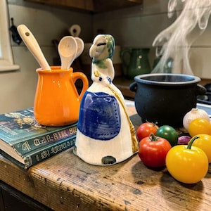 May include: A whimsical kitchen scene features a ceramic witch figurine with a blue apron, a cauldron, and fresh produce. An orange pitcher holds wooden spoons, and a book rests nearby. The scene evokes a cozy, rustic aesthetic.