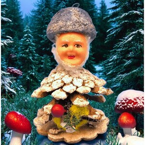 Puede incluir: Un adorno navideño caprichoso con forma de gnomo de seta. El gnomo tiene una cara sonriente, un gorro de seta gris y un cuerpo de piña. El adorno está ambientado en un fondo de bosque nevado con setas rojas.