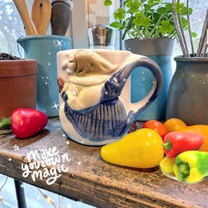 May include: A ceramic mug featuring a three-dimensional sculpted figure of a witch in a cream-colored hood and blue dress. The mug has a curved handle and a white base. The text "make your own magic" is written in white cursive on the surface.