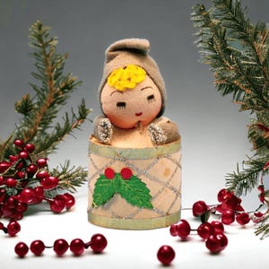 May include: A vintage Christmas ornament featuring a doll-like figure with a tan hood and a yellow flower on its head. The doll is inside a cylindrical container decorated with a green holly leaf and red berries. The ornament is surrounded by evergreen branches and red berry garlands.