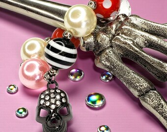 May include: A silver-toned skeleton hand holds a collection of decorative beads and a skull charm. The beads are various colors and patterns, including pearls, red with white polka dots, and black and white stripes. A black skull charm with rhinestone accents hangs from the beads.