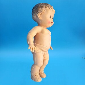 Puede incluir: Una mu&ntilde;eca beb&eacute; vintage con un tono de piel claro, de pie sobre un fondo azul. La mu&ntilde;eca tiene cabello rizado, mejillas rosadas y lleva un pa&ntilde;al y zapatos. Los brazos de la mu&ntilde;eca est&aacute;n ligeramente extendidos.