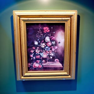 May include: A framed floral still life painting. The artwork features a vase overflowing with pink, white, and blue flowers. The painting is set against a dark background and framed in a gold-colored wooden frame. The artwork is mounted on a teal wall.