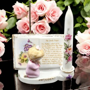 May include: A porcelain religious figurine set featuring a kneeling child, a candle, and a book with floral accents. The candle and book are white with pink and purple floral designs. The text "The Lord's Prayer" is visible.