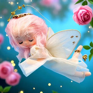 May include: A whimsical angel doll with pink hair, closed eyes, and a gold halo. The doll wears a white dress and has delicate white wings with gold accents. The background features pink roses and a soft blue hue.