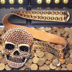 May include: A gold-colored belt with a skull-shaped buckle covered in rhinestones. The belt has a studded design and a leather strap. The skull's eyes are black. The belt is displayed on a pile of gold coins.