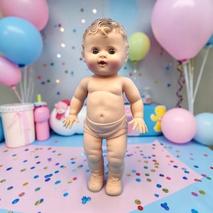 Puede incluir: Una mu&ntilde;eca beb&eacute; vintage con tono de piel neutro, que lleva pa&ntilde;ales y zapatos, de pie sobre una superficie rosa salpicada de confeti. La mu&ntilde;eca tiene ojos marrones y cabello rizado. Globos en rosa, azul y verde en el fondo.