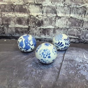 May include: Three ceramic spheres with a white base and blue floral patterns. The spheres vary in their specific designs, with some featuring larger floral motifs and others more intricate, smaller patterns. The spheres are arranged on a dark surface against a brick wall.