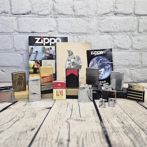 May include: A selection of Zippo lighters in diverse styles and colours, including gold, silver, and black. Some lighters have unique patterns and graphics, alongside Zippo promotional materials. The lighters are displayed on a wooden surface.