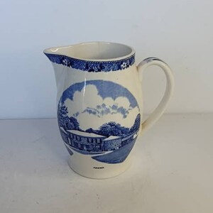 May include: A white ceramic pitcher with a blue and white design. The pitcher features a scene of a house with a blue sky and clouds. The words "AGNES" are printed on the bottom of the pitcher.