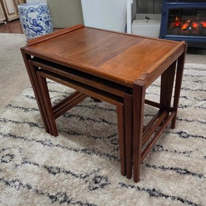 May include: A set of three nested tables crafted from medium-toned wood. The tables feature rectangular tops with slightly raised edges and straight legs, designed to slide neatly underneath one another for compact storage and versatile use.