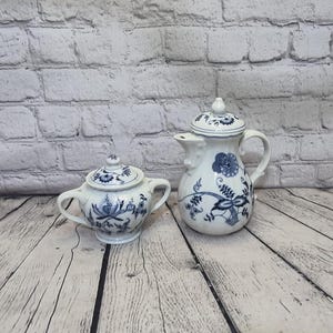 May include: A white porcelain creamer and sugar bowl set with blue floral designs. The creamer has a spout and a handle. The sugar bowl has a lid and two handles.