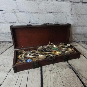 May include: A collection of antique spoons of various designs and sizes, displayed inside an open, dark brown wooden chest. The spoons are made of silver and gold-toned metals, with decorative handles and intricate details. The chest is lined with a brown fabric.