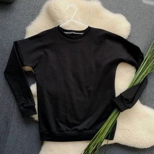 Könnte beinhalten: Ein schwarzer Langarm-Pullover mit Rundhalsausschnitt. Der Pullover hängt an einem weißen Kleiderbügel und liegt auf einer weißen, flauschigen Oberfläche.