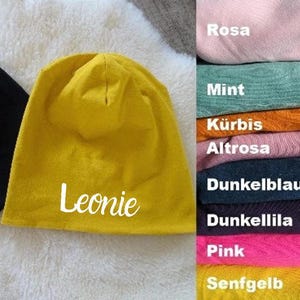 Könnte beinhalten: Eine gelbe Mütze mit dem Namen "Leonie" bestickt auf der Vorderseite. Die Mütze wird vor einem Hintergrund aus Stoffmustern in verschiedenen Farben gezeigt.