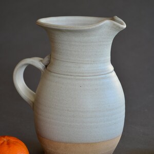 May include: A handmade, light blue ceramic pitcher with a handle and a spout. The pitcher has a wide base and a narrower neck. The bottom of the pitcher is a light brown color. The pitcher is likely used for serving beverages.
