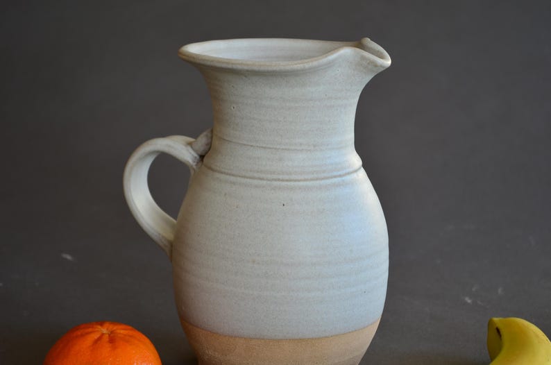 May include: A handmade, light blue ceramic pitcher with a handle and a spout. The pitcher has a wide base and a narrower neck. The bottom of the pitcher is a light brown color. The pitcher is likely used for serving beverages.