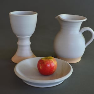 May include: A set of light gray ceramic tableware consisting of a tall stemmed goblet, a small pitcher with a handle, and a shallow bowl. A single red apple with yellow highlights sits inside the bowl. Each piece has a natural tan base.