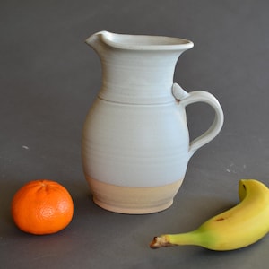 May include: A ceramic pitcher with a light blue glaze and a tan base, featuring a handle and a spout. An orange tangerine and a yellow banana are placed near the pitcher on a gray surface.