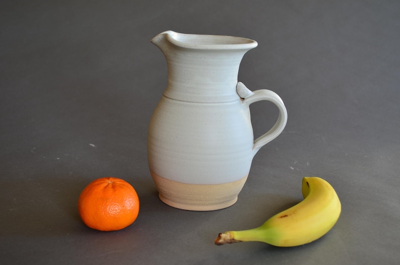 May include: A ceramic pitcher with a light blue glaze and a tan base, featuring a handle and a spout. An orange tangerine and a yellow banana are placed near the pitcher on a gray surface.