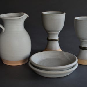 May include: A collection of handmade ceramic tableware in a neutral color palette. The set includes a pitcher, two goblets, and two small bowls. The pieces have a light gray glaze with a natural, unglazed base. The goblets have a dark green band.