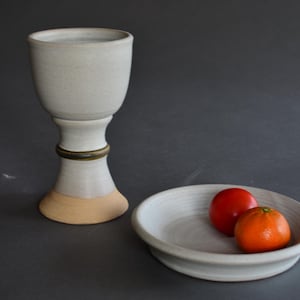 May include: A ceramic chalice with a light gray glaze, a brass band, and a tan base, next to a small, round, light gray ceramic bowl holding a red tomato and an orange tangerine. The objects are on a dark gray surface.