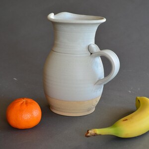May include: A handmade, light gray ceramic pitcher with a handle and a wide spout. The base is a light tan color. An orange tangerine and a yellow banana are next to the pitcher.