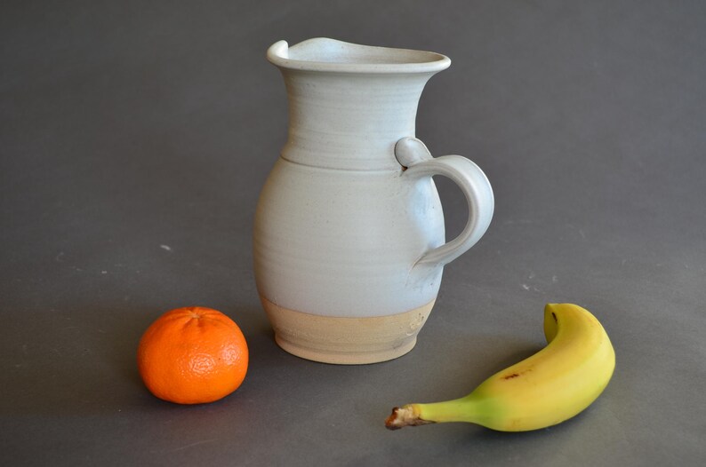 May include: A handmade, light gray ceramic pitcher with a handle and a wide spout. The base is a light tan color. An orange tangerine and a yellow banana are next to the pitcher.