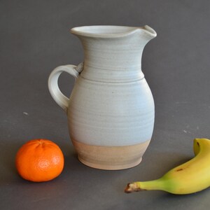 May include: A handmade ceramic pitcher with a light blue glaze and a tan base. An orange tangerine and a yellow banana are next to the pitcher. The pitcher has a handle and a spout.