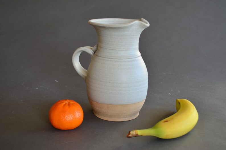 May include: A handmade ceramic pitcher with a light blue glaze and a tan base. An orange tangerine and a yellow banana are next to the pitcher. The pitcher has a handle and a spout.
