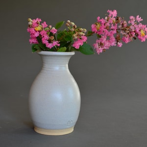 May include: A white ceramic vase with a light beige base holds vibrant pink flowers. The vase has a rounded body and a slightly flared rim. The flowers have small, clustered blooms and green leaves.