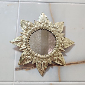 Puede incluir: Espejo decorativo en forma de estrella con un marco de metal dorado. El marco presenta un diseño floral detallado con un espejo circular en el centro. El espejo está montado sobre una superficie de baldosas de color claro.