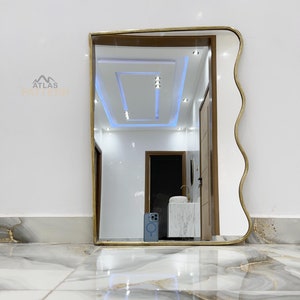 May include: A gold-framed mirror with a wavy edge, reflecting a white ceiling with recessed lighting and a hallway with a white door and a white sink.