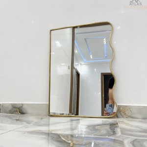 May include: A gold-framed wavy mirror with a white ceiling and white walls reflected in the glass. The mirror is sitting on a white and gray marble floor.