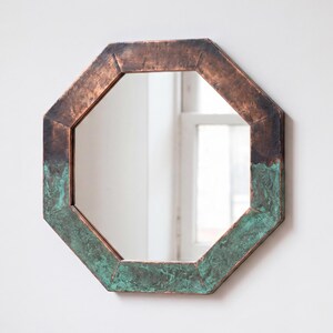 May include: An octagonal wall mirror featuring a frame with a dual-tone finish. The upper portion of the frame displays a smooth, dark metallic copper tone, while the lower section exhibits a textured, oxidised verdigris green patina finish.