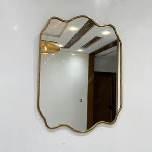 May include: A gold-framed mirror with a wavy, scalloped edge. The mirror reflects a room with a white ceiling and a brown door.