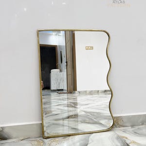 May include: A wavy mirror with a gold frame, reflecting a bathroom interior. The mirror is on a white marble floor.