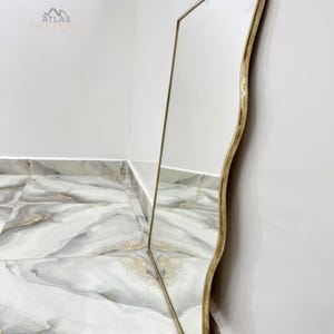 May include: A gold-coloured, wavy-edged mirror with a reflective surface. The mirror is leaning against a white wall and a marble-patterned floor.