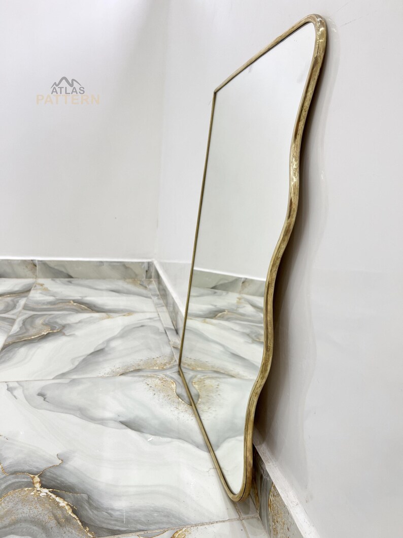 May include: A gold-coloured, wavy-edged mirror with a reflective surface. The mirror is leaning against a white wall and a marble-patterned floor.
