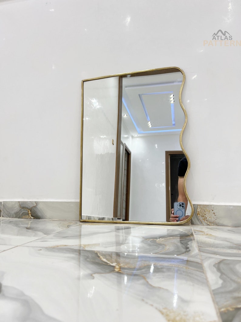 May include: A gold-framed wavy mirror with a white ceiling and white walls reflected in the glass. The mirror is sitting on a white and grey marble floor.