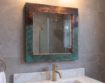 Puede incluir: Un espejo de pared cuadrado con un marco ancho y plano de acabado oxidado y desgastado, que transita de un tono metálico marrón oscuro en la parte superior a una vibrante pátina verde azulada texturizada en los lados y la base.