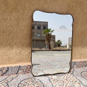 May include: A gold-framed mirror with a wavy, ornate design. The mirror reflects a building with a palm tree in front of it. The text "ATLAS PATTERN" is visible in the reflection.