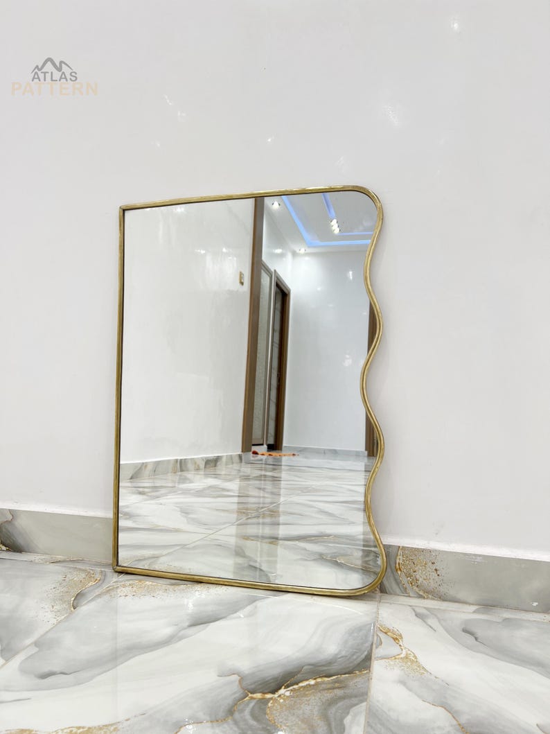 May include: A gold-framed mirror with a wavy edge. The mirror is reflecting a hallway with white walls and tiled floors.