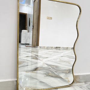 May include: A wavy mirror with a gold frame and a slightly curved edge. The mirror is reflecting a tiled floor and a door.