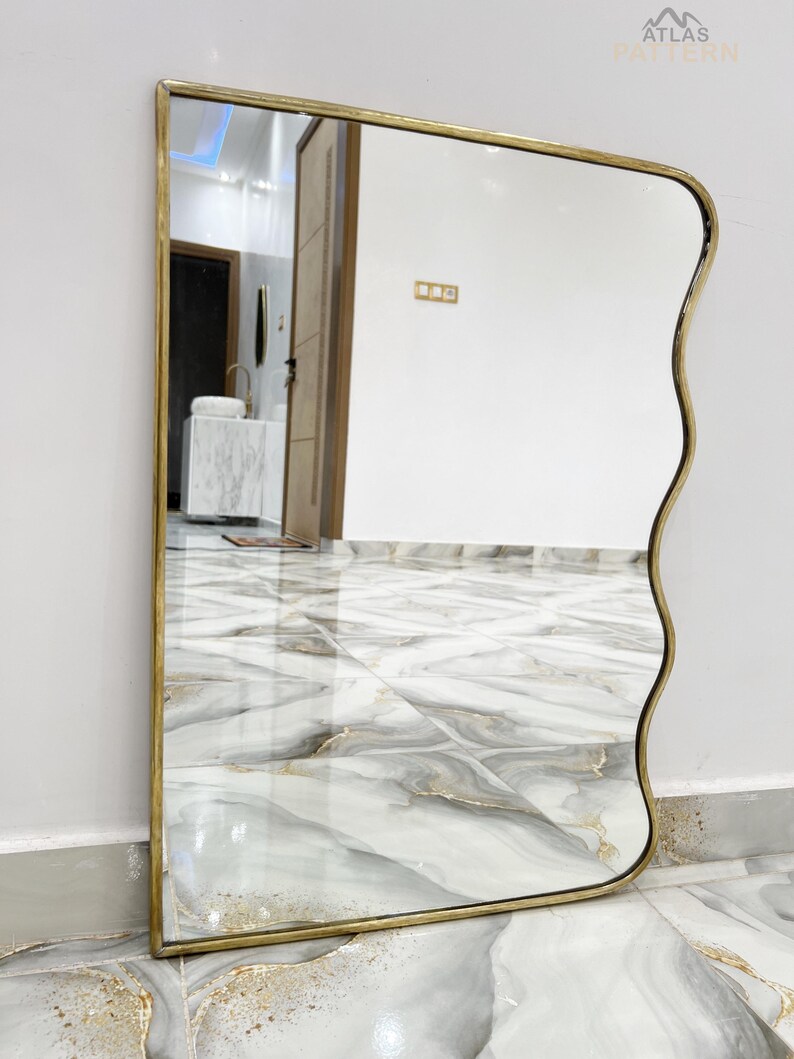 May include: A wavy mirror with a gold frame and a slightly curved edge. The mirror is reflecting a tiled floor and a door.