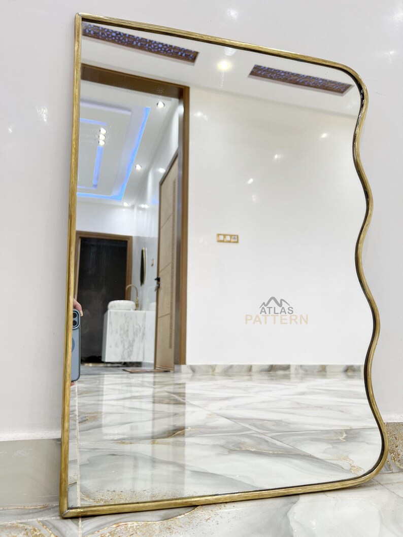 May include: A gold-framed mirror with a wavy edge. The mirror is reflecting a room with white walls and a tiled floor.