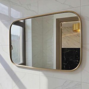 May include: A rectangular wall mirror with rounded corners and a thin, metallic gold-toned frame. The mirror is mounted on a wall covered in white marble-patterned tiles, reflecting a window, a doorway, and a gold light fixture.