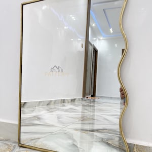May include: A gold-framed mirror with a wavy edge. The mirror is reflecting a hallway with a tiled floor.
