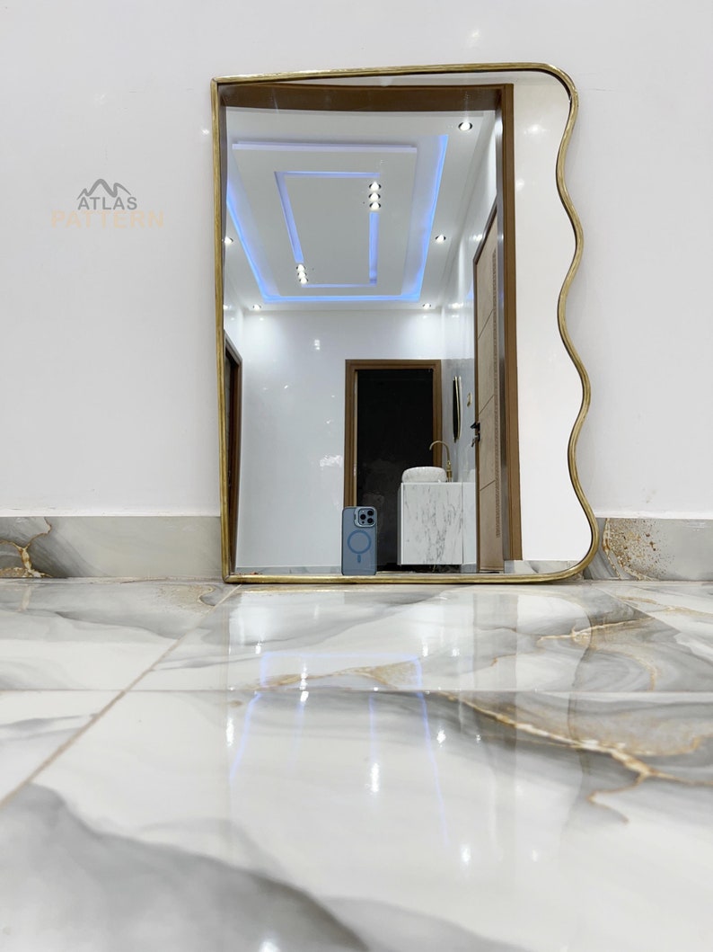 May include: A gold-framed mirror with a wavy edge, reflecting a white ceiling with recessed lighting and a hallway with a white door and a white sink.