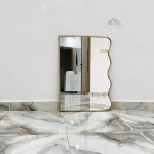 May include: A wavy mirror with a gold frame against a white background. The mirror is reflecting a white sink and a white countertop.